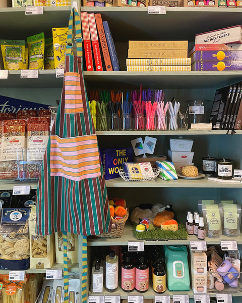 Specialty-goods shop shelves filled with food, cookbooks, colorful pens, a striped apron, pet products, toys, and candles.