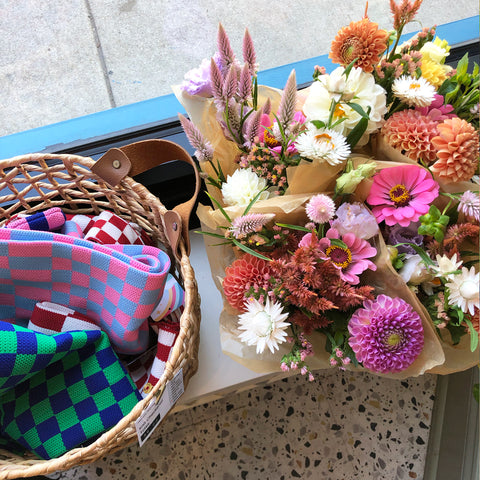 Colorful flower bouquets beside a wicker basket filled with bright checkerboard-patterned tote bags.