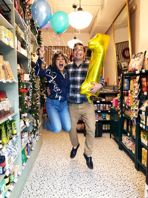 Two adults celebrate in a decorated shop with balloons, ribbons, and a large gold number one balloon.