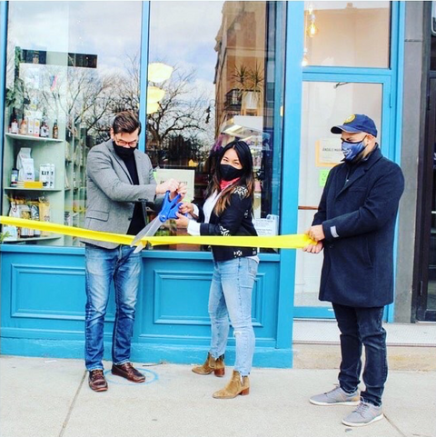 Three masked people stand outside Ándale Market in Andersonville. One cuts a yellow ribbon with large scissors at a ribbon cutting ceremony.