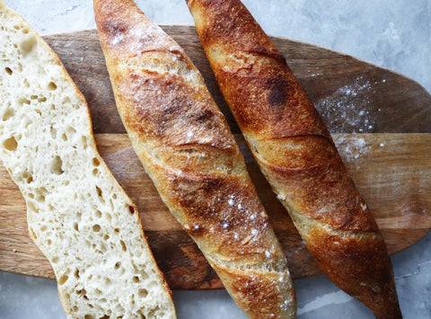 Two golden-brown baguettes and a cut section of airy bread on a wooden cutting board.
