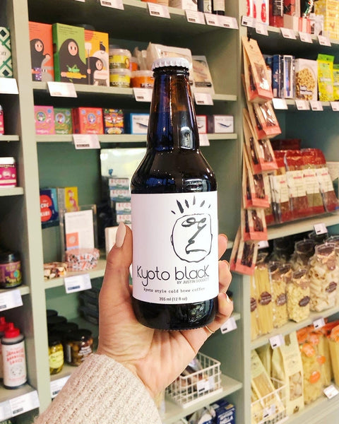 A hand holds a brown bottle of Kyoto Black cold brew coffee against green retail shelves.