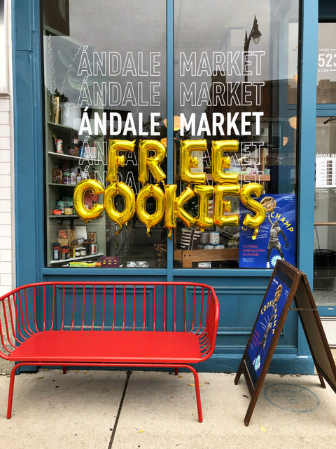 Ándale Market storefront with gold “FREE COOKIES” balloons, a cookie-event poster, and a red outdoor bench.