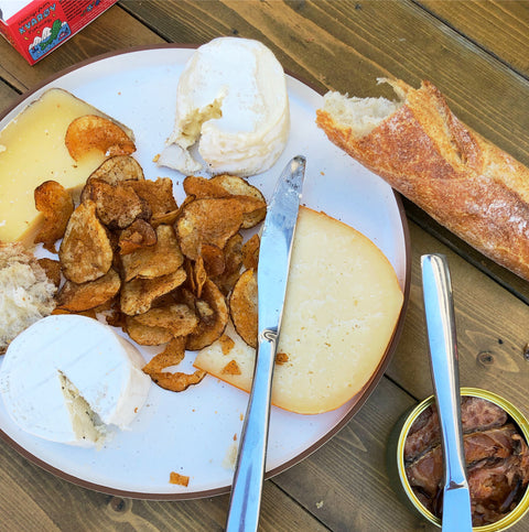 A serving plate with cheese, chips, crusty bread, tinned fish, and serving utensils on a wooden table.