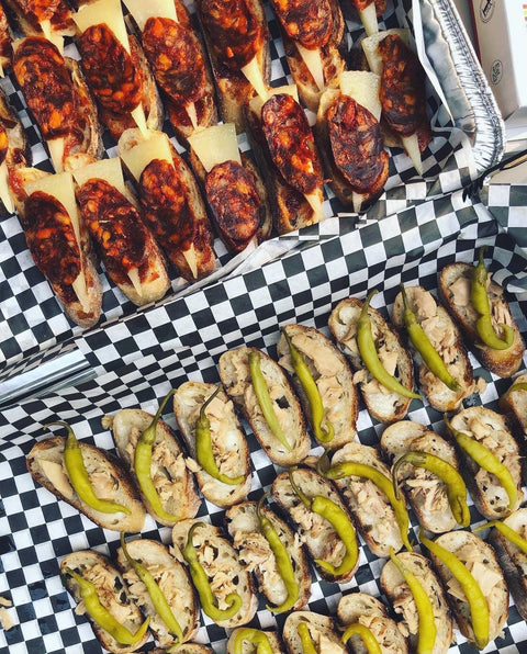 Overhead view of assorted open-faced pinxtos on checkered paper, with meat, cheese, fish, and peppers.