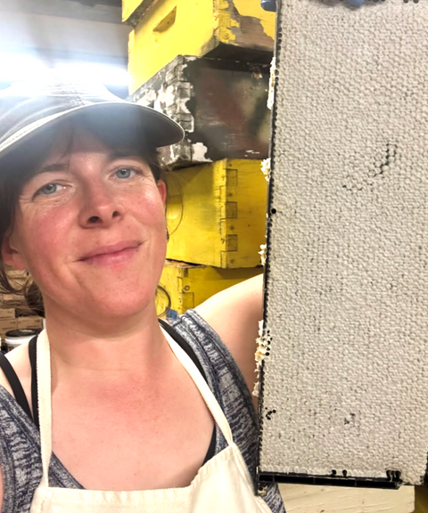 Bike A Bee founder in a baseball cap and apron posing beside a honeycomb panel.
