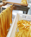 Yellow dried pasta strips hang from wooden rods next to a white plastic basket filled with loose yellow pasta in a production facility.