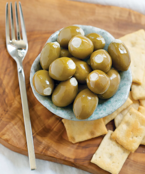 Glossy green olives in a small bowl on a wooden board, with square crackers and a silver fork nearby.