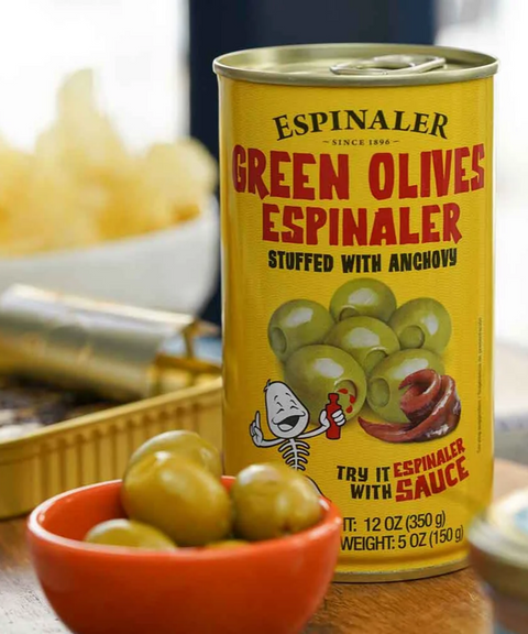 A yellow can of Espinaler Green Olives stuffed with anchovy on a wooden surface with a small orange bowl of green olives.