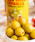 Glossy green olives in a small white bowl with a blurred yellow can of Espinaler anchovy stuffed olives.