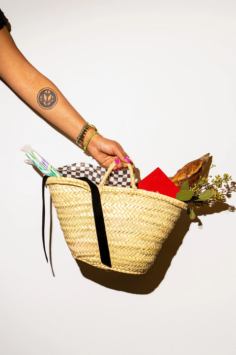 Hand holding a woven straw tote filled with colorful items, bread, and leafy flowers against a white background.