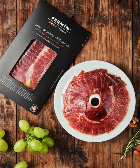 Fermin 100% Bellota cured ham sliced on a plate next to its black package on a wooden surface.