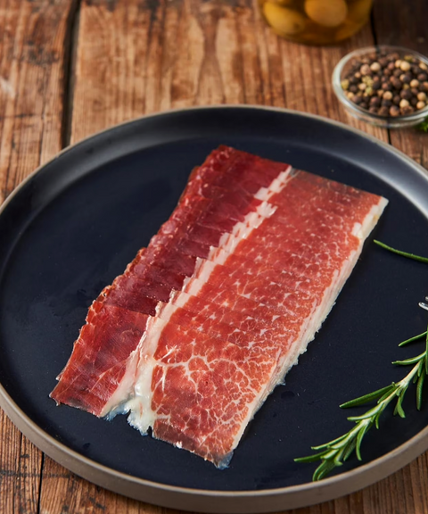 Slices of cured ham with marbled red and white fat, arranged on a dark round plate.