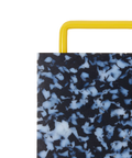 Close-up of a rectangular plastic cutting board with a black and white marbled pattern and yellow metal handle, shown on a plain white background.