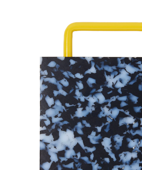 Close-up of a rectangular plastic cutting board with a black and white marbled pattern and yellow metal handle, shown on a plain white background.