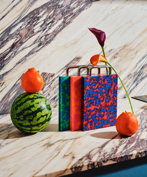 Three colorful patterned cutting boards on a marble surface with fruit props and a flower stem around them.