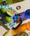A colorful speckled plastic cutting board with a black handle, styled on a wood table with limes and cocktails nearby.
