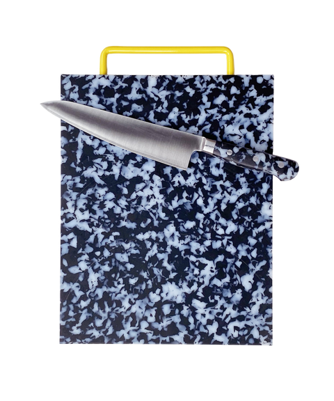 A rectangular kitchen cutting board with a black and white marbled pattern and a bright yellow handle, shown with a chef's knife laid diagonally across the top surface on a plain white background.
