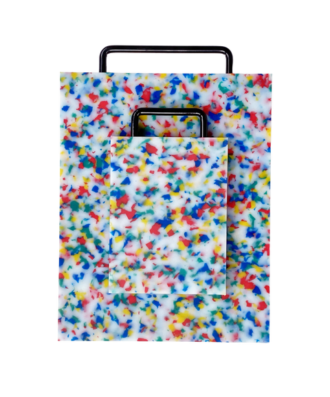 A rectangular plastic cutting board with a black metal handle, featuring a white base covered in dense multicolored confetti-like speckles in blue, red, yellow, and green, shown on a plain white background. There is a smaller matching version of the same cutting board on top of the larger one.