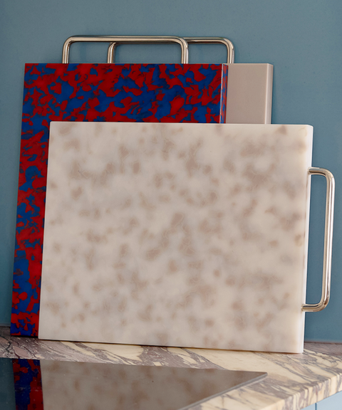 A marble kitchen cutting board with a white and grey marbled pattern and a silver metal handle, displayed on a marble countertop against a blue wall. Other cutting boards are stacked behind it.