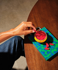 A hand holds a red candy over a small yellow bowl filled with red candy, placed on a colorful blue and green patterned cutting board on a wooden table.