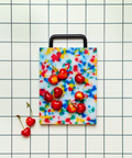 A plastic rectangular cutting board with a black handle, featuring a colorful confetti pattern on a white background. The cutting board is photographed from above on a white grid-patterned surface with loose cherries.
