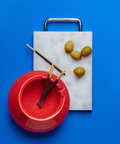 A rectangular plastic cutting board with a grey and white marbled pattern and silver chrome metal handle, styled on a bright blue background with a red speckled ceramic ashtray, olives, and thin biscuits dipped in chocolate.