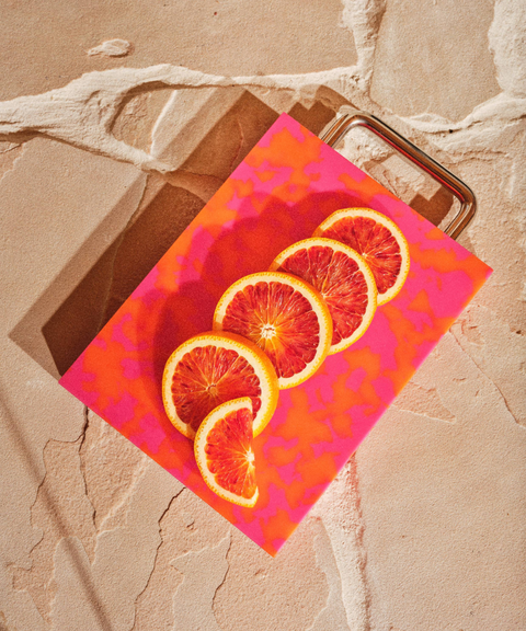 A rectangular plastic cutting board with a pink and orange marbled pattern and silver chrome metal handle, styled on a beige stone background and blood orange slices.