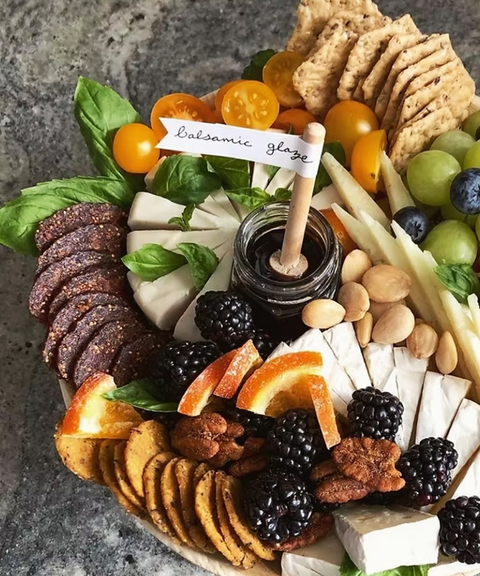 A beautifully arranged charcuterie board featuring an assortment of cheeses, crackers, fruits, nuts, Hellenic Farms fig salami, and a small jar of balsamic glaze with a wooden stick.