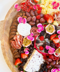 A close-up of a large wooden charcuterie board filled with cheeses, sliced meats, fruit, flowers, and Hellenic Farms Fig Salami.