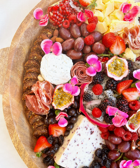 A close-up of a large wooden charcuterie board filled with cheeses, sliced meats, fruit, flowers, and Hellenic Farms Fig Salami.