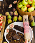 A bowl filled with sliced Hellenic Farm fig salami sits among fresh figs, apples and strawberries on a wooden surface.