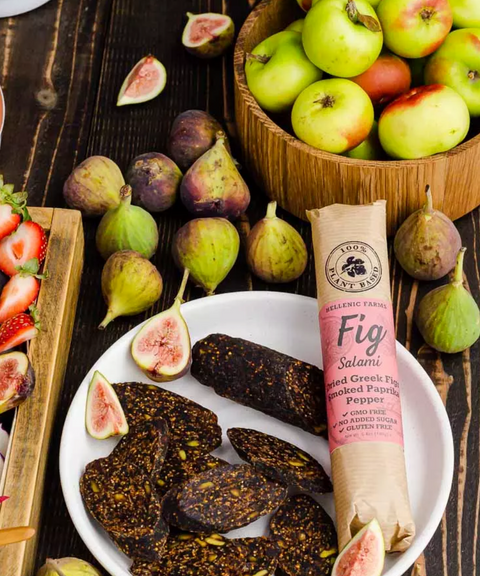 A bowl filled with sliced Hellenic Farm fig salami sits among fresh figs, apples and strawberries on a wooden surface.