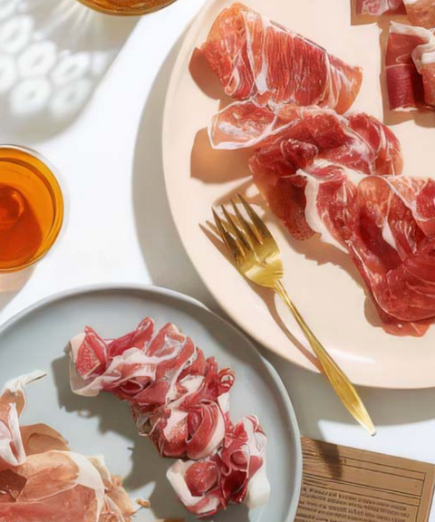 Thinly sliced prosciutto arranged on two plates, with a gold fork resting on the larger plate. 