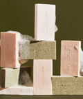 A stacked arrangement of Malin + Goetz soap bars in pale pink, speckled sage green, and cream, with lather around some bars.