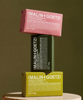 Three stacked Malin + Goetz bar soap packages in pink, dark green, and yellow-green displayed on a round pedestal.