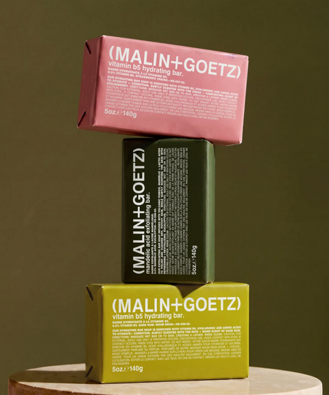 Three stacked Malin + Goetz bar soap packages in pink, dark green, and yellow-green displayed on a round pedestal.