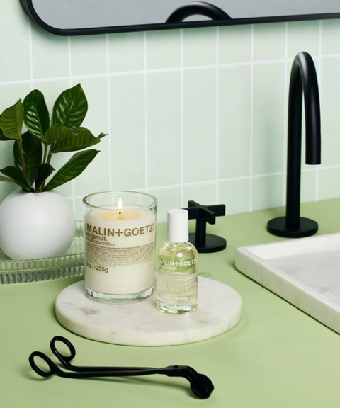 A modern bathroom countertop features a lit Malin + Goetz Beramot candle and a bottle of oil on a marble tray.