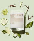 A Malin + Goetz Bergamot candle in a glass jar surrounded by lime and bergamot slices, green leaves, and twigs on a pastel green background.