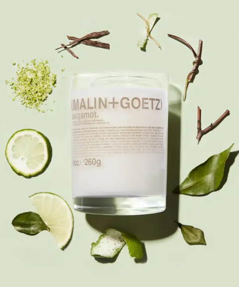 A Malin + Goetz Bergamot candle in a glass jar surrounded by lime and bergamot slices, green leaves, and twigs on a pastel green background.