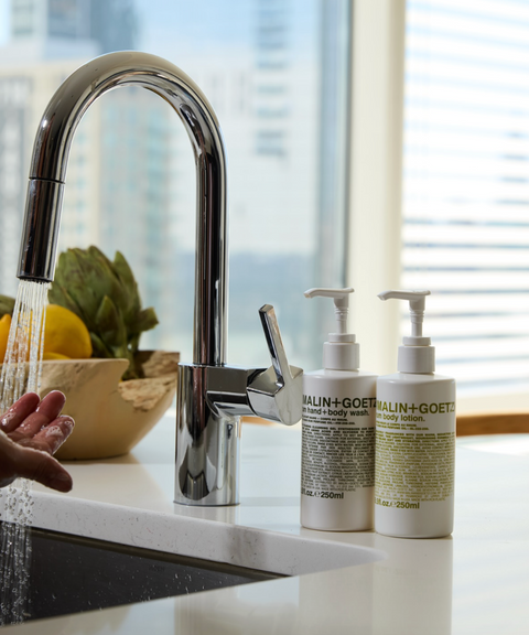 Running water from a chrome kitchen faucet over a hand beside two Malin+Goetz pump bottles.