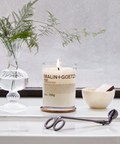 A lit Malin+Goetz Bergamot candle on a wooden coaster, flanked by a glass vase with green sprigs and candle scissors, with a small bowl nearby.