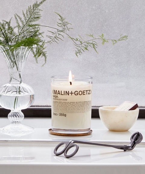 A lit Malin+Goetz Bergamot candle on a wooden coaster, flanked by a glass vase with green sprigs and candle scissors, with a small bowl nearby.