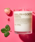 A Malin+Goetz Strawberry candle in a clear glass jar next to a white strawberry and leaves.