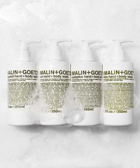 Four white Malin+Goetz hand and body wash pump bottles arranged in a row against a foamy white background.