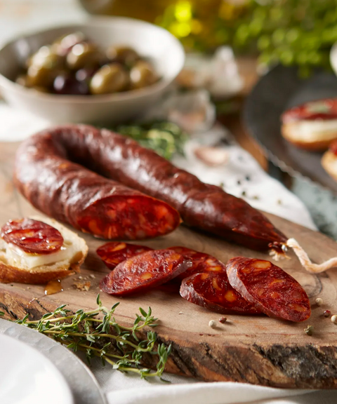 Sliced Palacios chorizo arranged on a rustic wooden board with bread, herbs and olives.