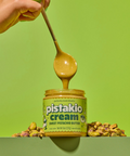 Open jar of Pistakio pistachio cream with a spoonful of thick spread dripping into it, surrounded by pistachios.