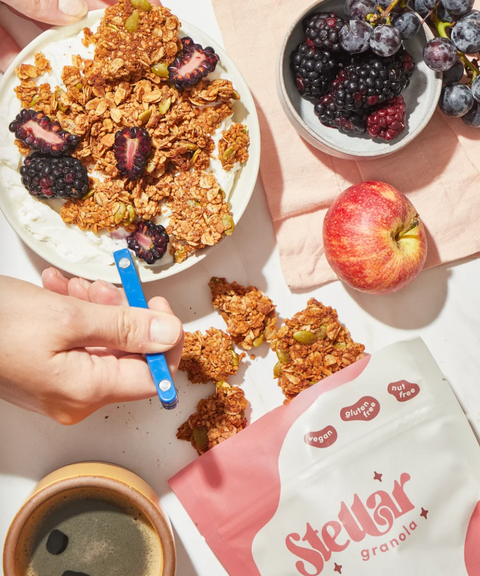 A hand holds a spoon in a bowl with granola, alongside fresh berries, an apple and a bag of Stellar Granola.