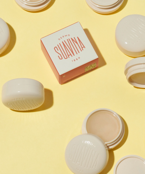 A square box of Suavina original lip balm surrounded by cream-colored round lip balm pots on a yellow background.