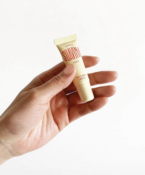 A hand holding a cream-colored tube of Suavinia original lip balm.